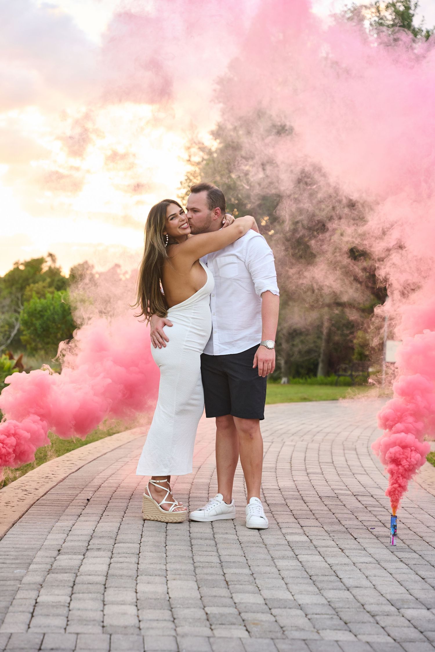 Couple embracing during a gender reveal with pink smoke bombs on a brick path at sunset.