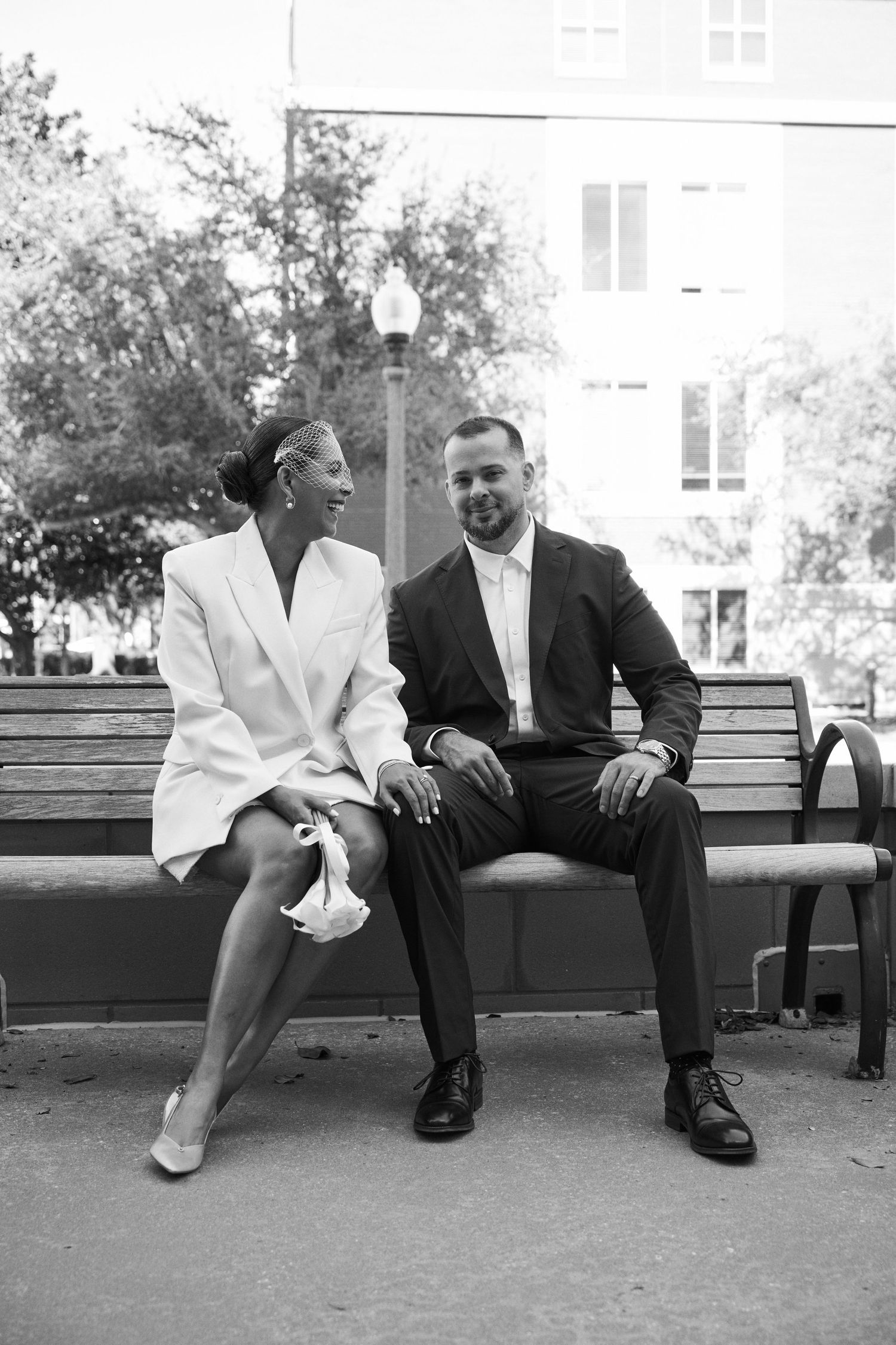 Black and white photo of newlyweds sitting on park bench laughing together after courthouse wedding.