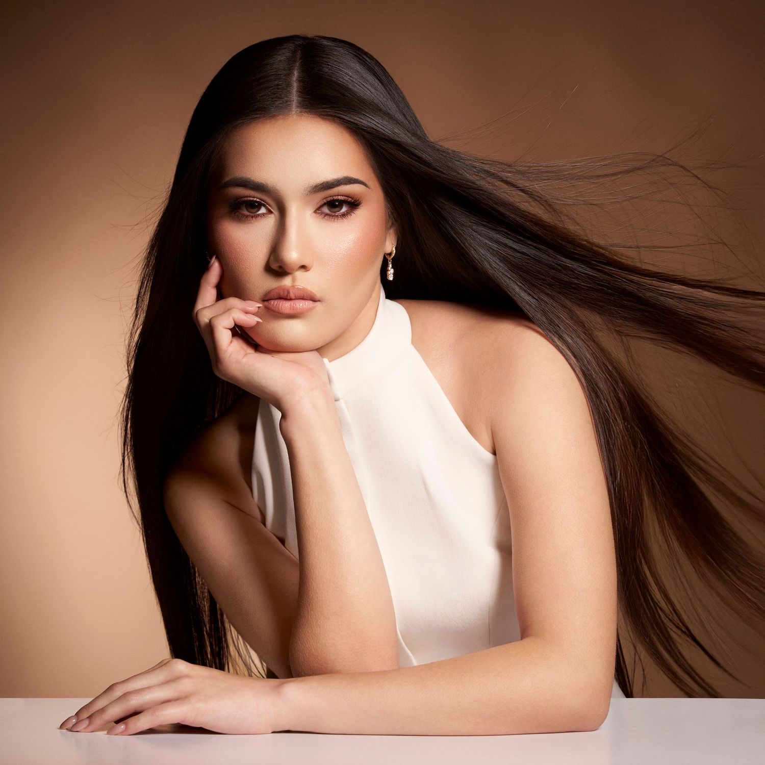 A young woman with long flowing dark hair poses elegantly in a white halter top against a warm beige background.