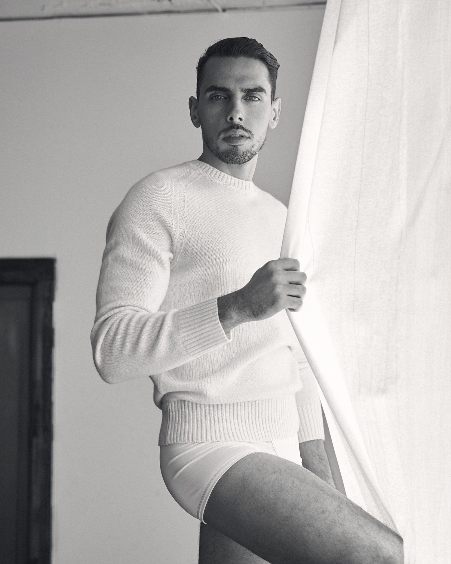Black and white photo of a man in white sweater and briefs holding a sheer curtain indoors.