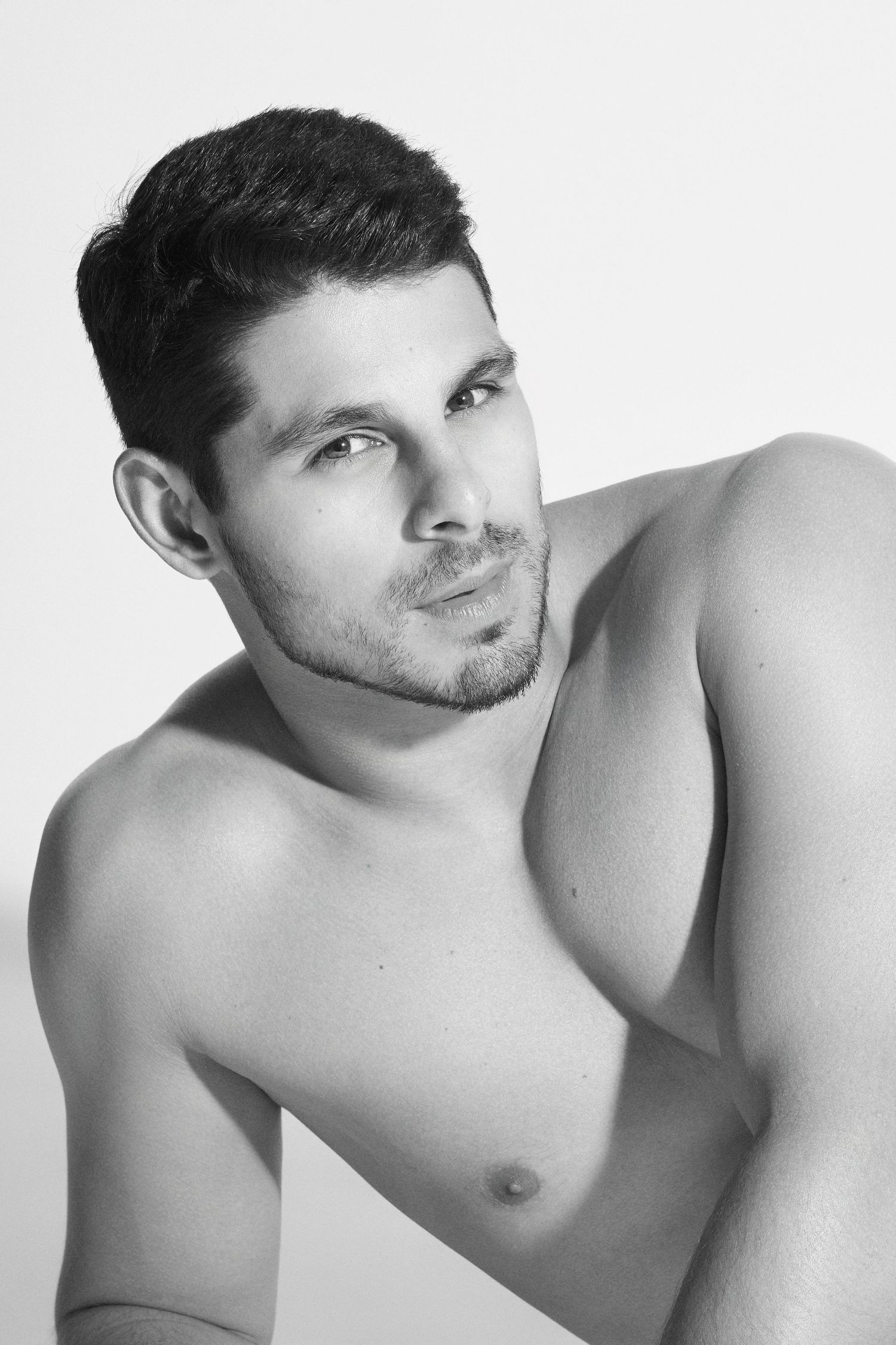 Black and white portrait of a muscular shirtless young man with dark hair and stubble posing against white background.