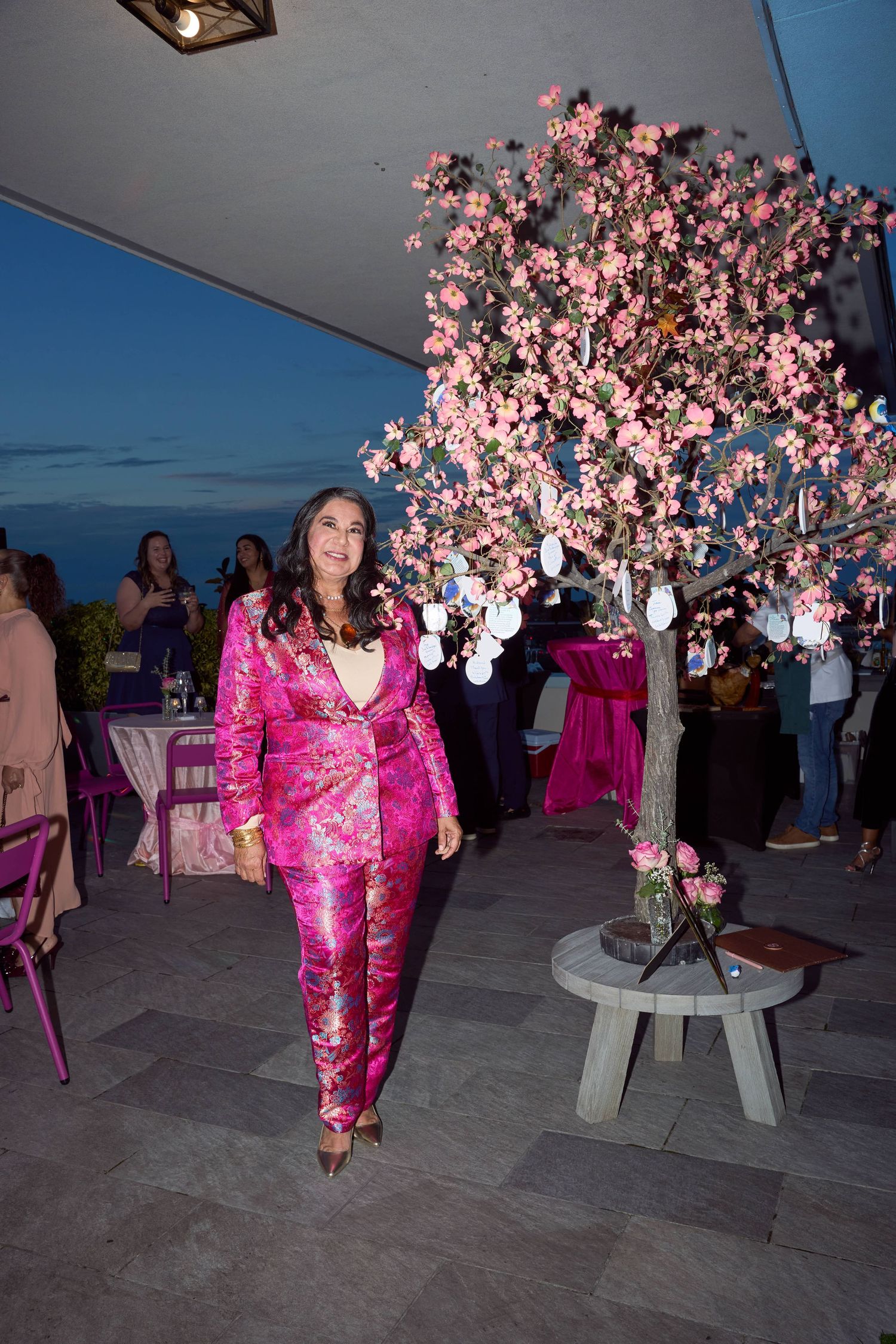Woman in pink floral jumpsuit poses beside a blooming cherry blossom tree at an elegant evening outdoor event.