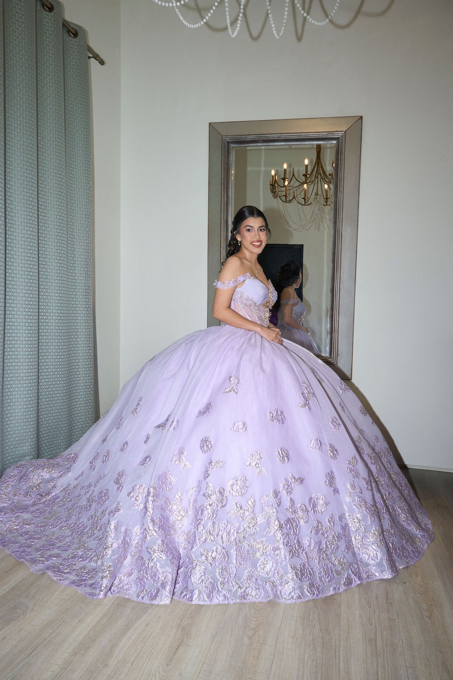 Young woman in lavender off-shoulder ball gown with floral embroidery posing in front of large mirror.