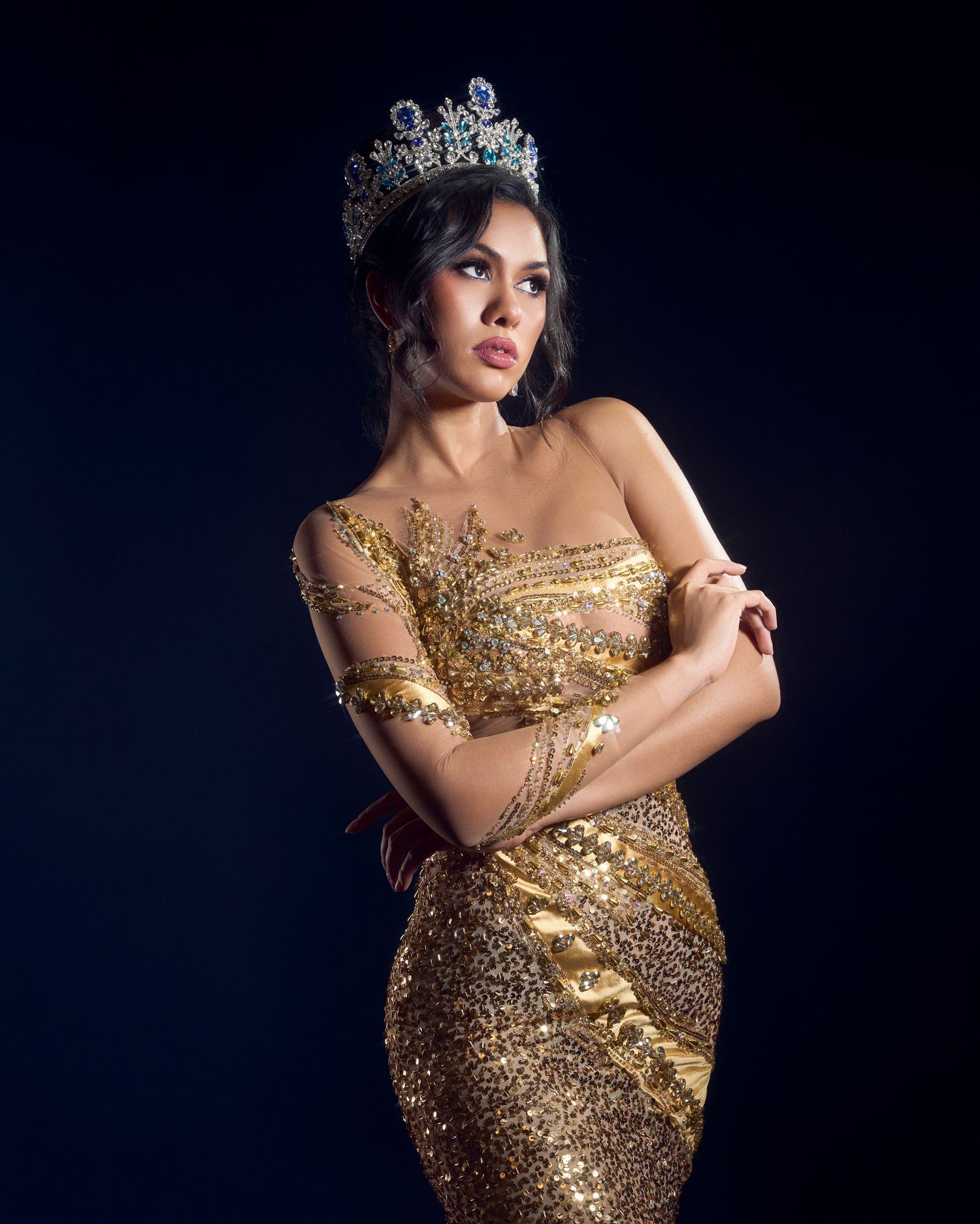 Woman wearing sparkling gold gown and diamond tiara crown posing elegantly against dark studio background.