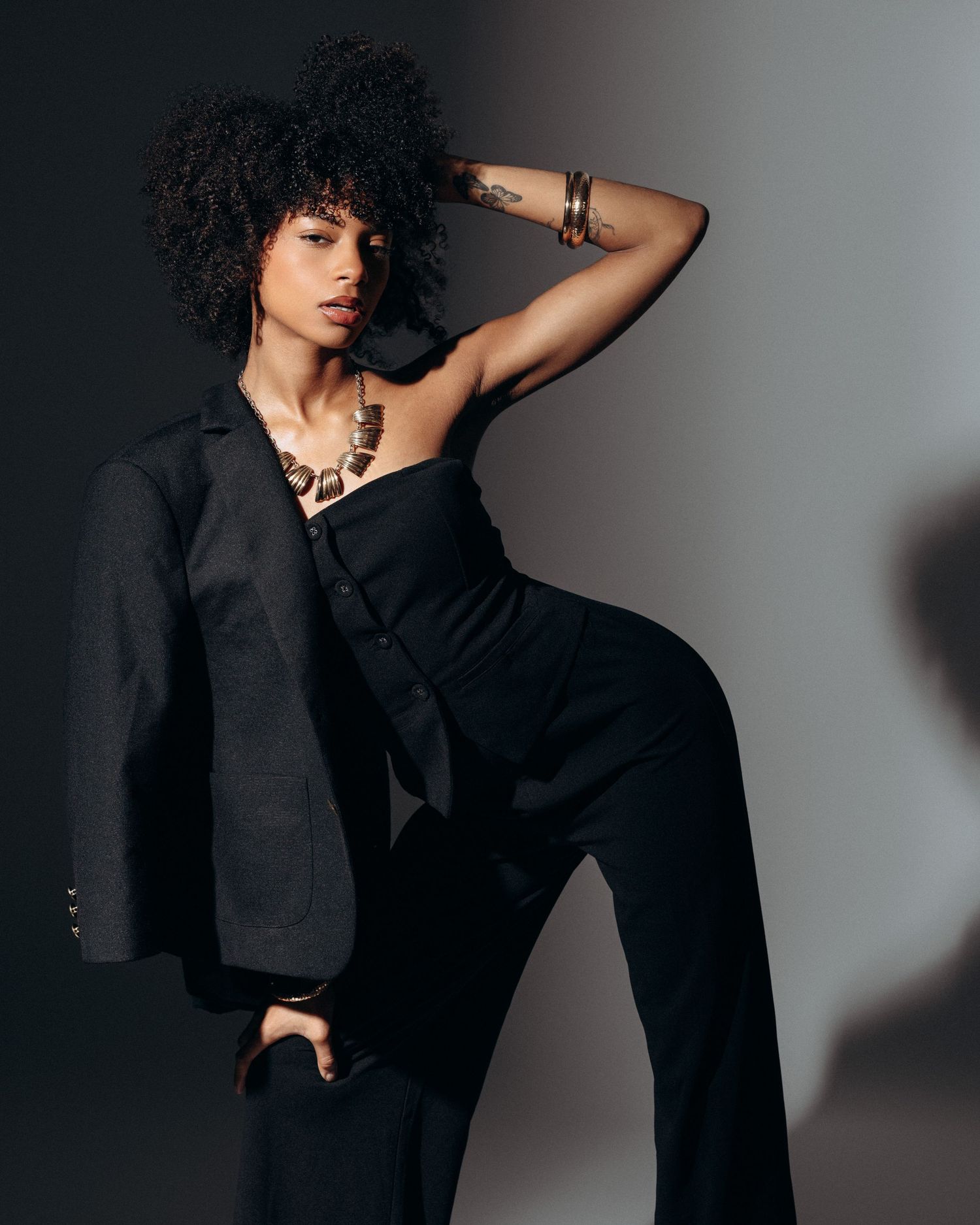 Fashion portrait of a female model in a black suit posing against a minimalist studio background with dramatic lighting.