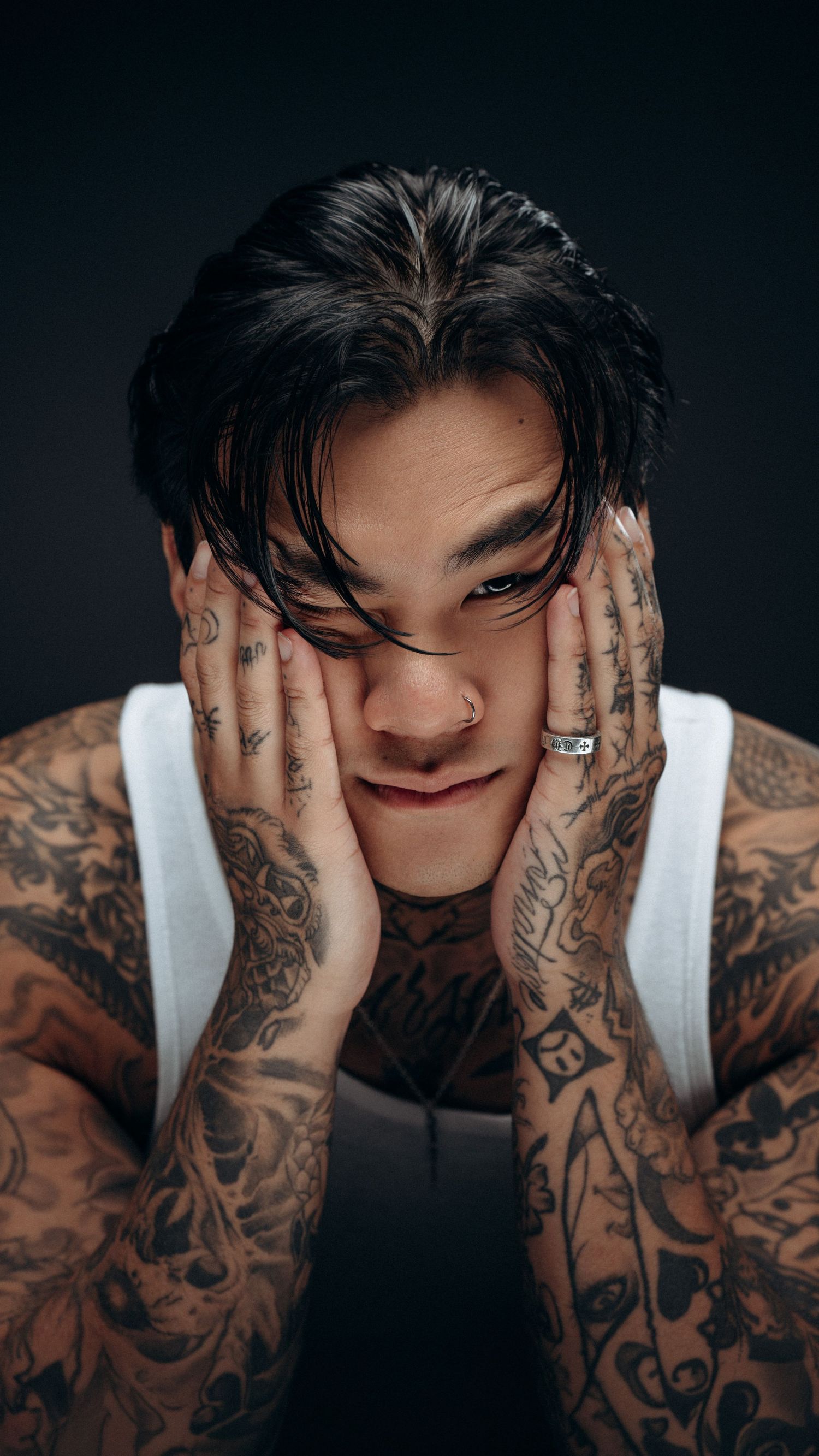 Expressive portrait of a tattooed male model with hands on face, in white tank, photographed in a dark studio setting in Montreal, Quebec.