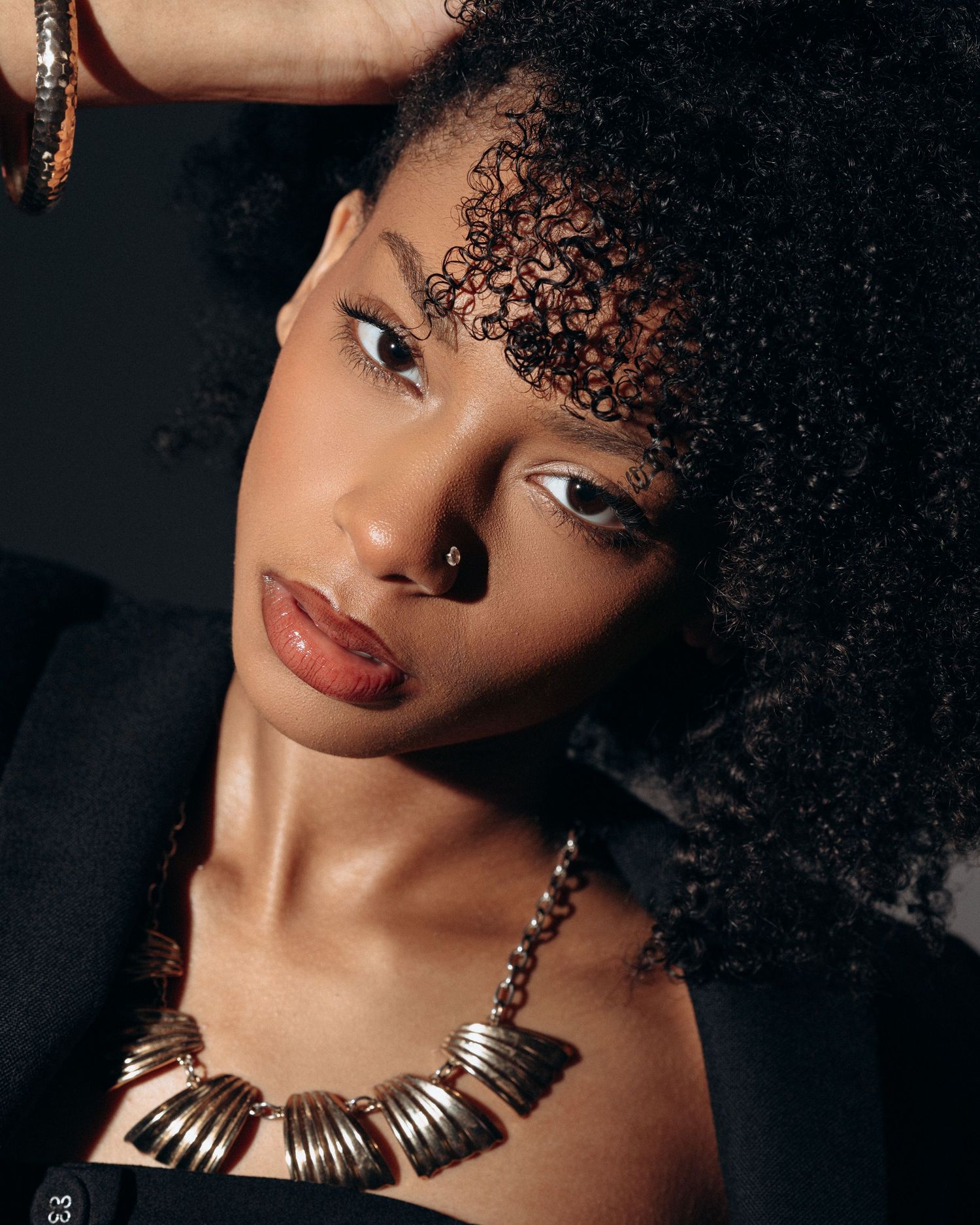 Close-up portrait of a female model with natural curls and statement jewelry in a softly lit studio setting.