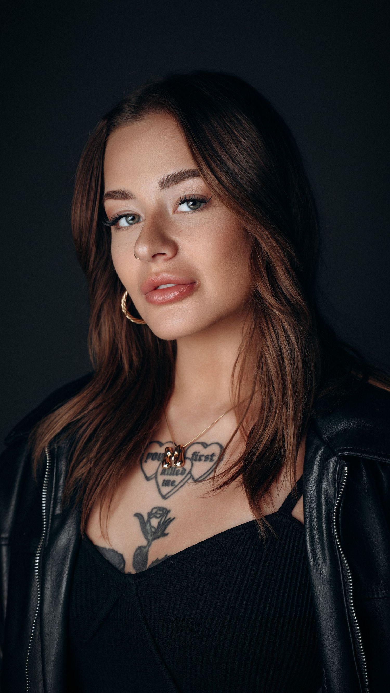 Moody portrait of a female model in a black outfit with subtle jewelry, tattoos, captured in a dark studio setting by Montreal photographer.