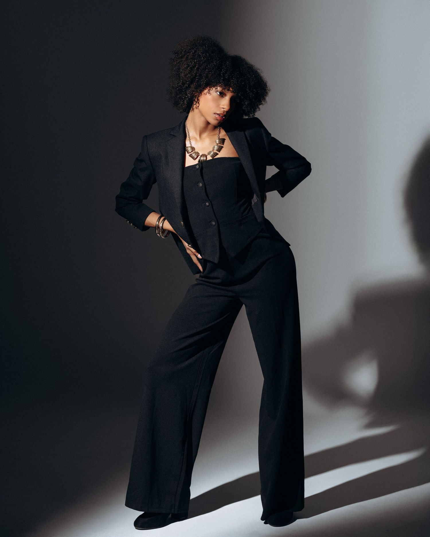 Full-body fashion portrait of a female model in a black outfit with directional lighting and a minimalist background.
