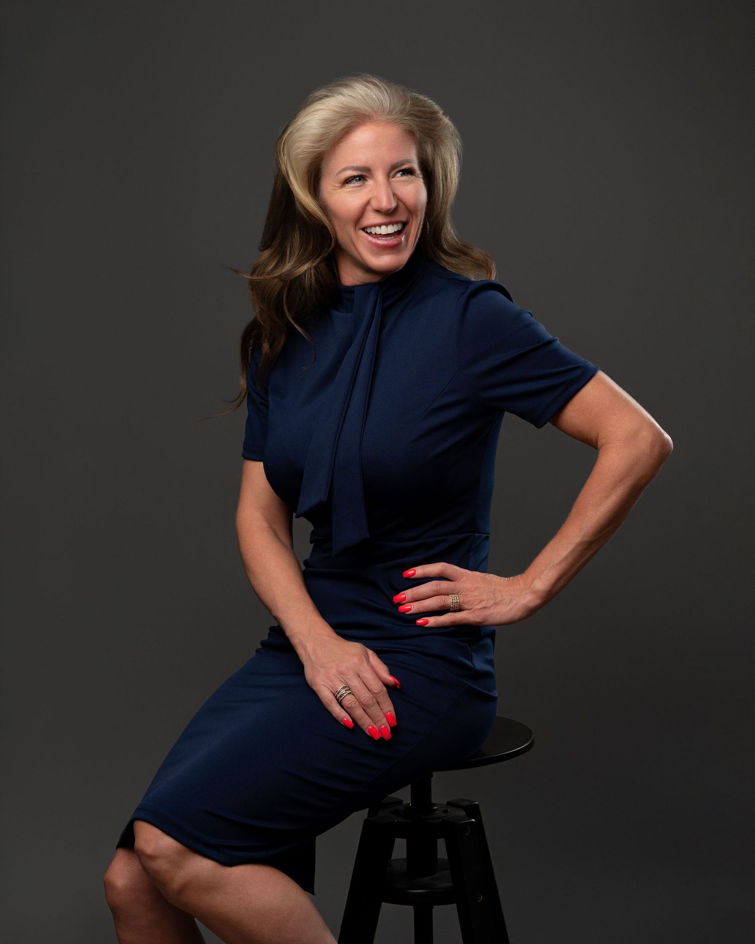 Elegant portrait of a woman seated on a stool in a navy dress with soft, refined studio lighting.