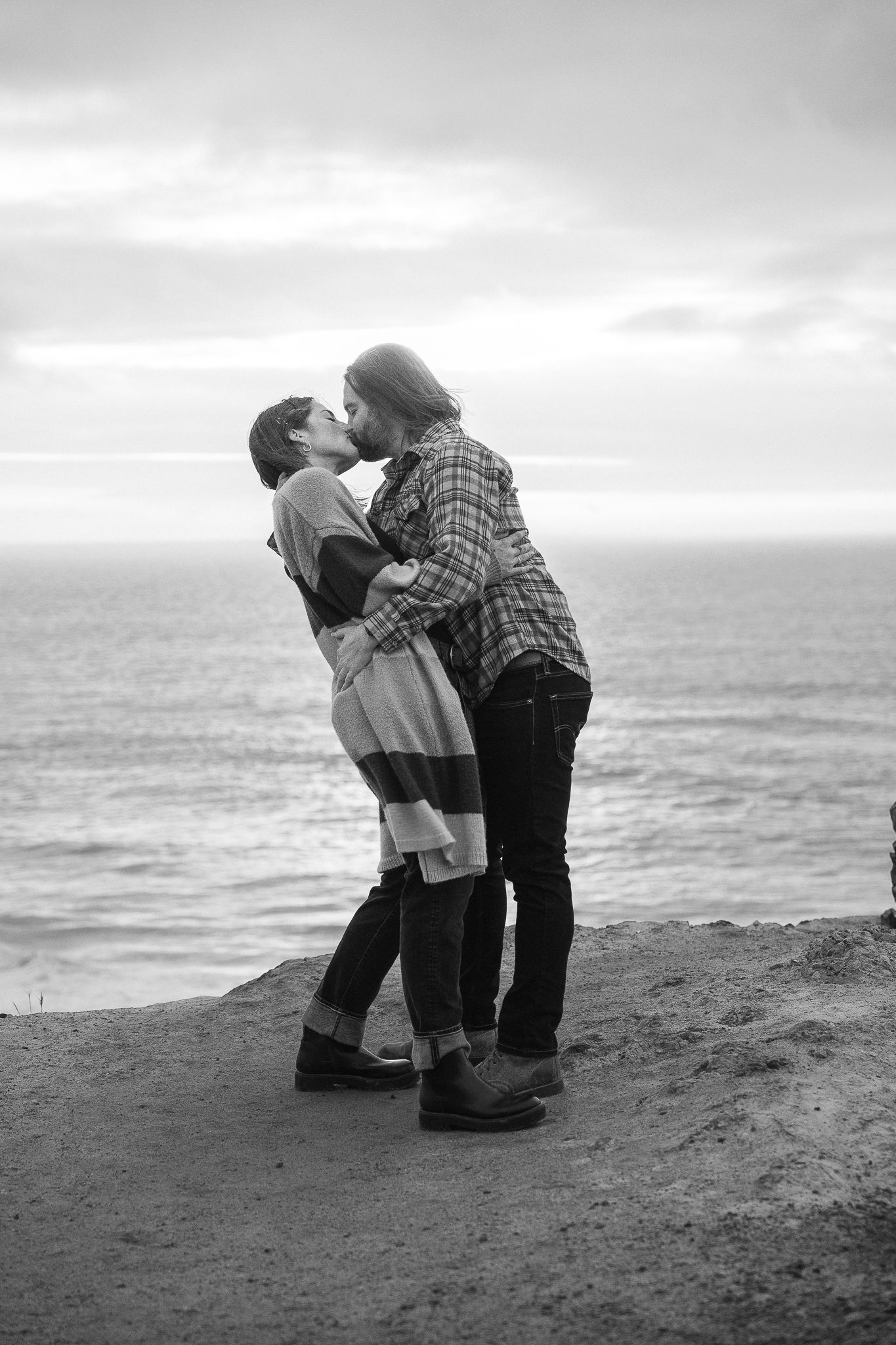 Black and white photograph of a couple in the quiet, breathless moment of a proposal with the ocean behind them