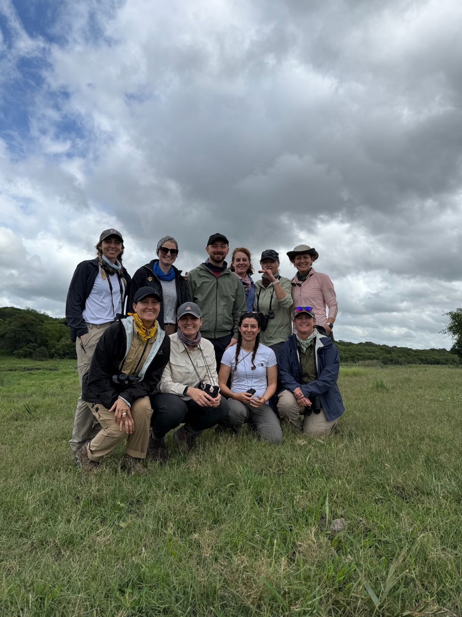 A group of people sitting and standing in a grassy field in South Africa with a cloudy sky.