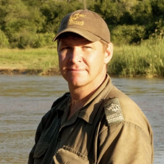 A man in a tan hat and jacket stands near a body of water in a natural setting.