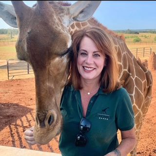 A smiling woman in a green uniform stands next to a giraffe in an outdoor setting.