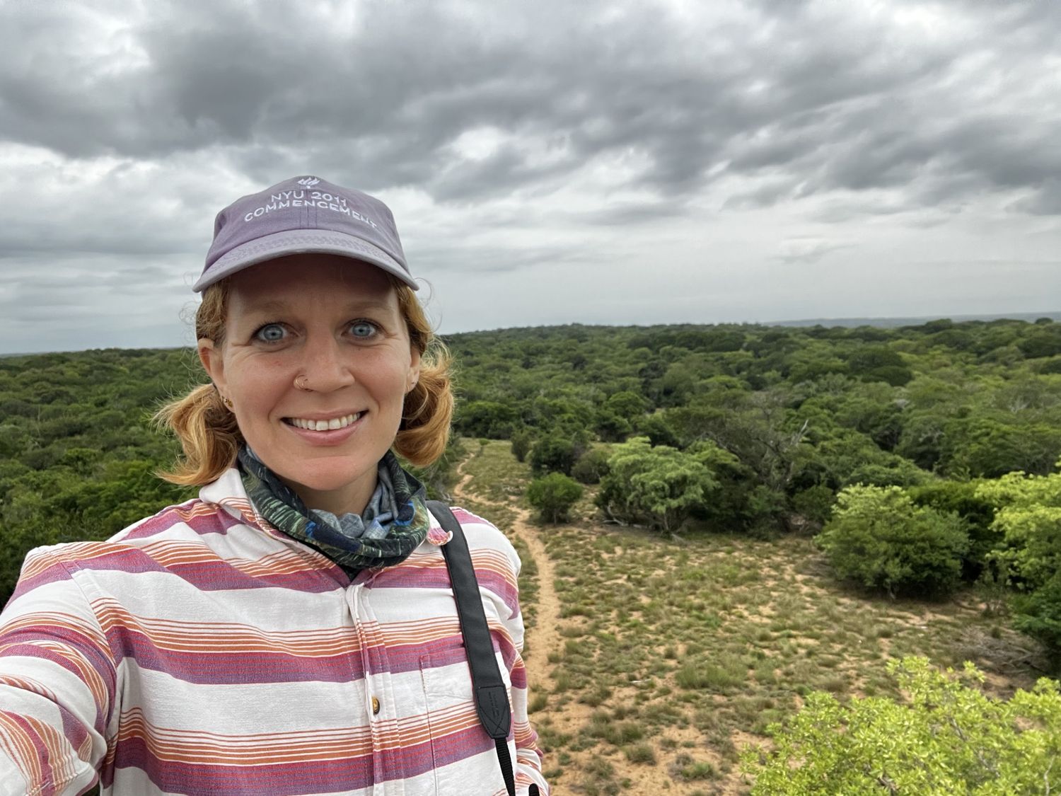 A woman wearing a striped shirt and hat stands on a hill overlooking a lush, green landscape in South Africa.