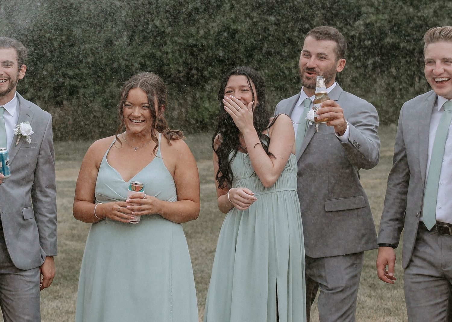 Wedding party laughing and celebrating outdoors in sage green bridesmaid dresses and grey suits.