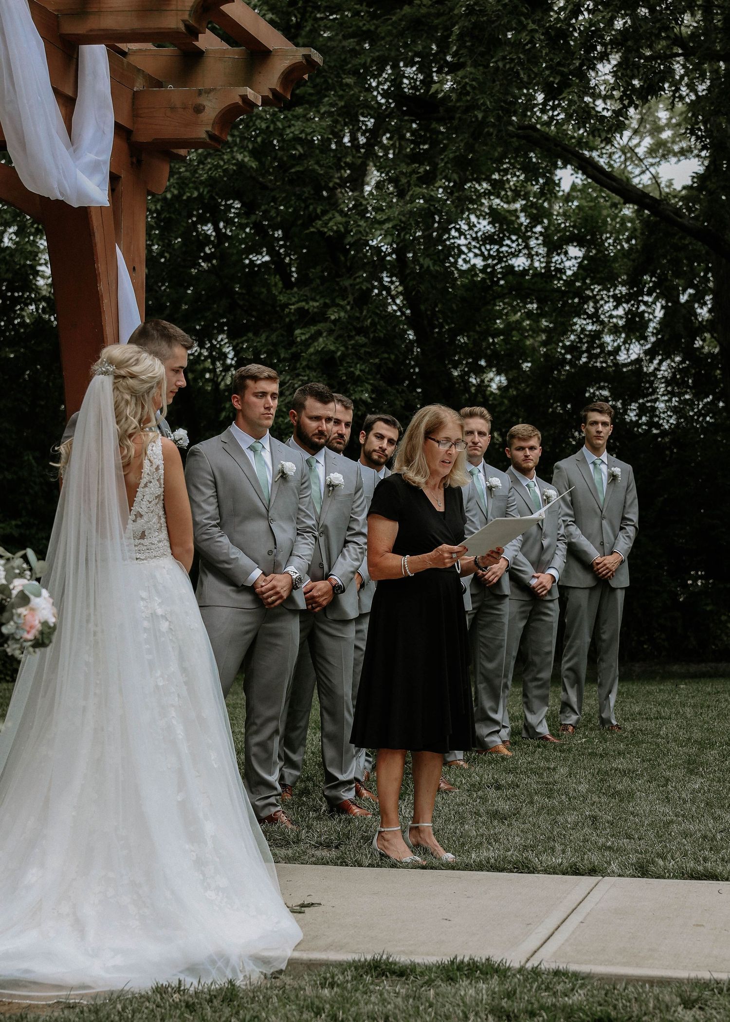 Outdoor wedding ceremony with bride in white gown, officiant in black dress, and groomsmen in gray suits under wooden arbor.
