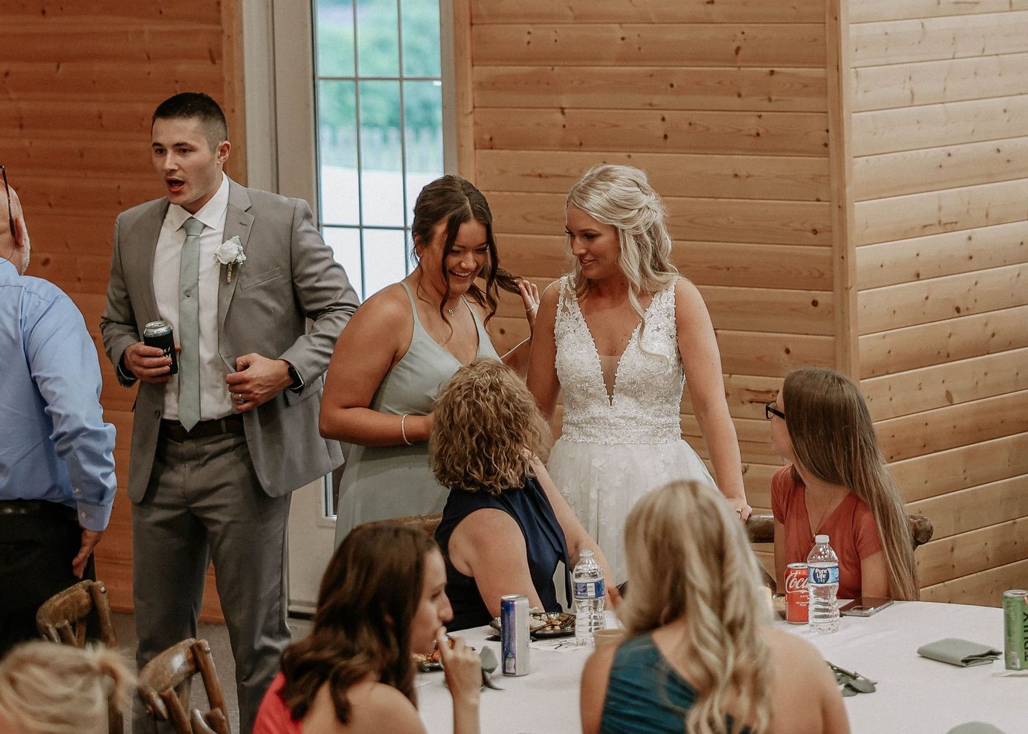 Bride in white lace gown laughing with wedding party guests at a rustic wooden barn reception.