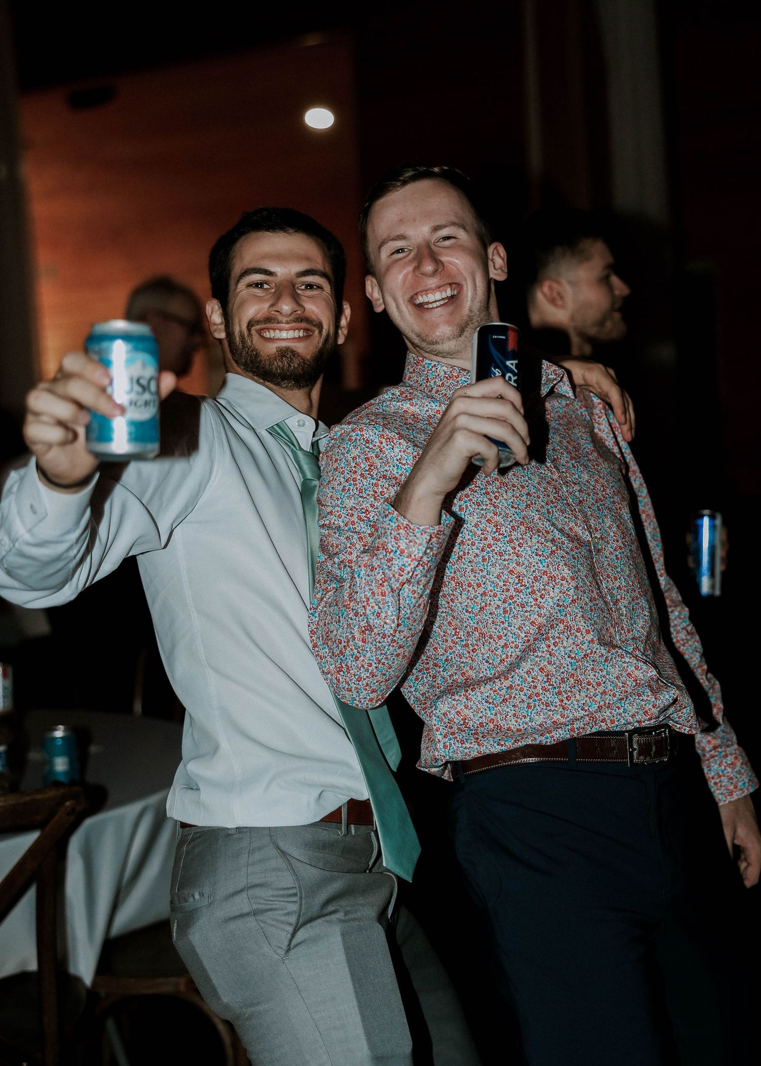 Two smiling men in dress shirts raising beer cans at a lively wedding reception party.