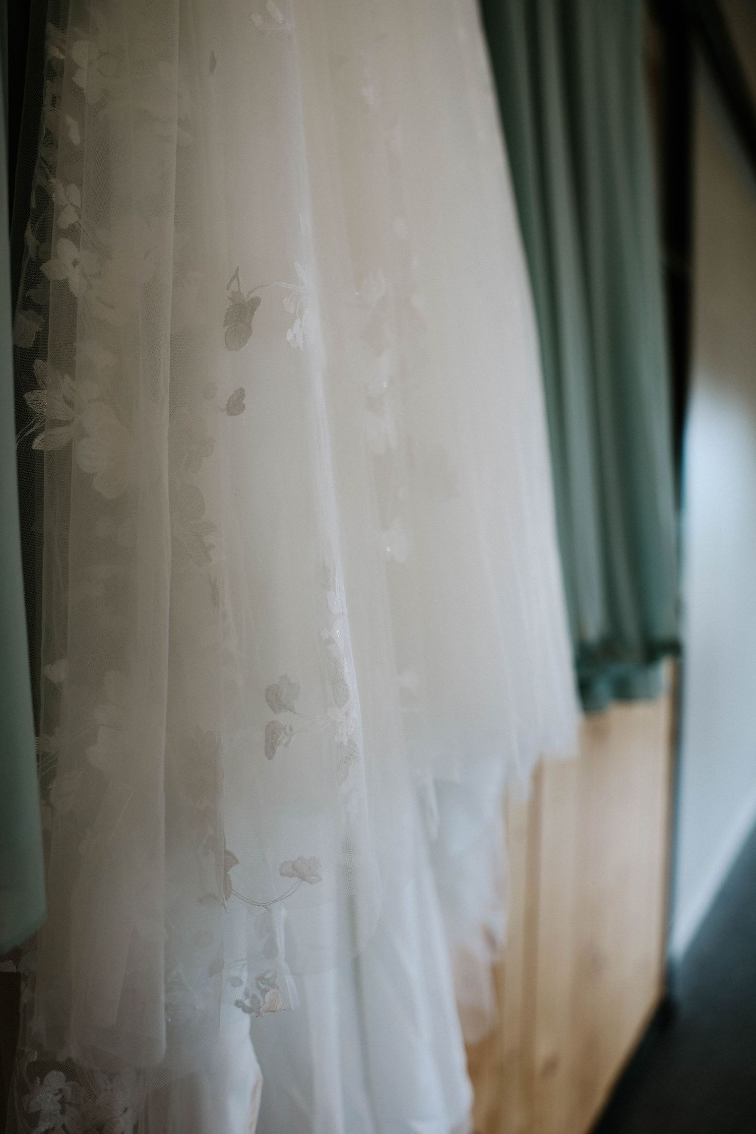 Close-up of a white floral lace wedding dress hanging near green curtains in soft natural light.