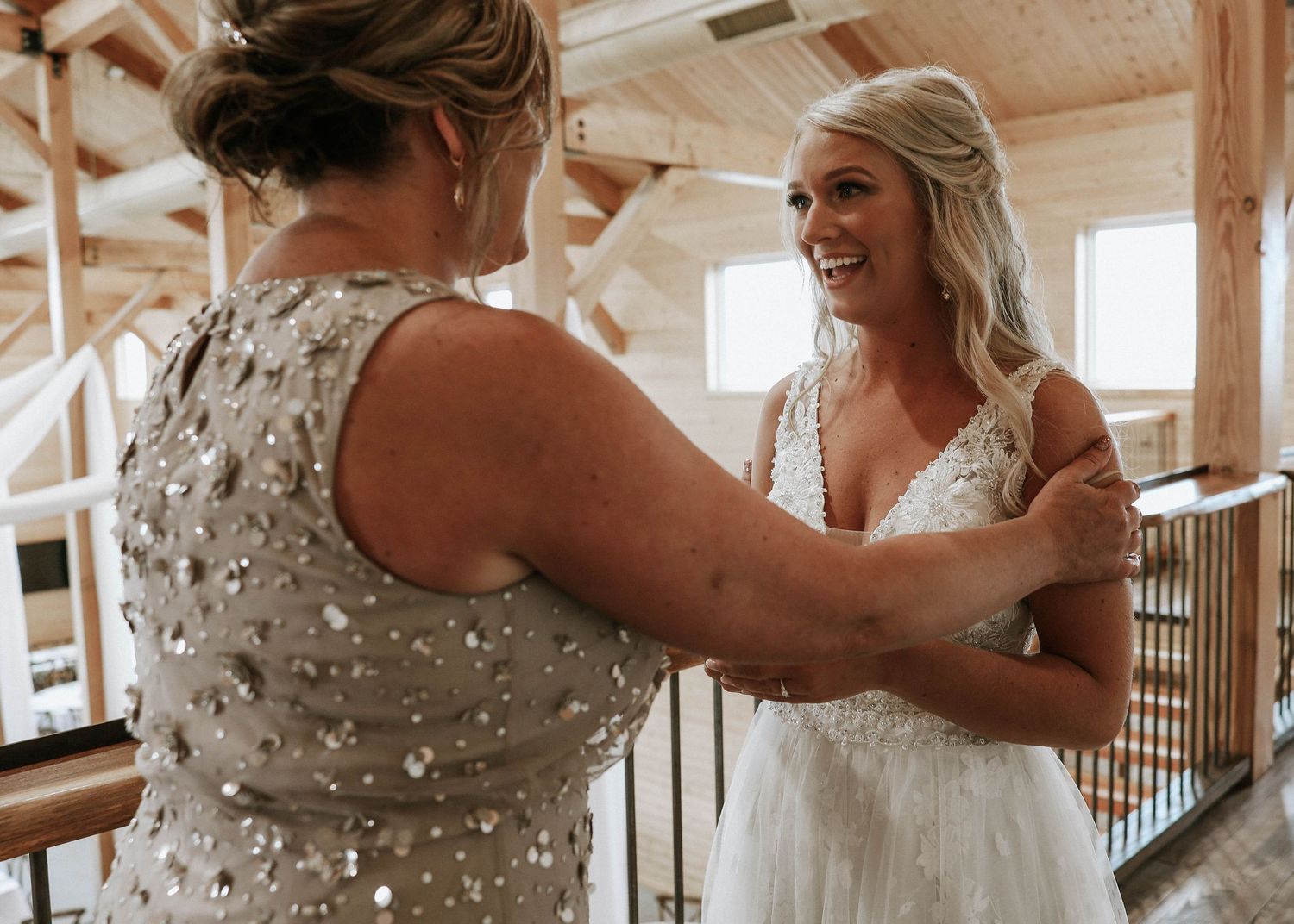 Bride smiling as woman in sequined dress helps fasten her lace wedding gown in rustic barn venue.