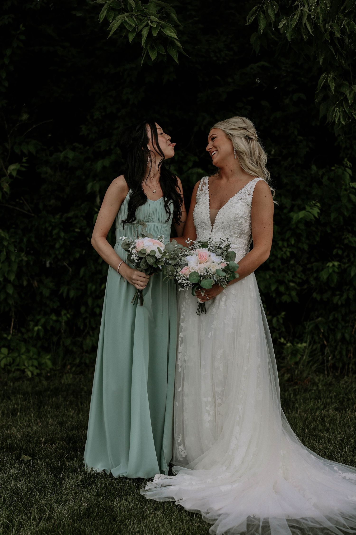 Bride in lace gown and bridesmaid in sage green dress sharing a joyful moment outdoors.