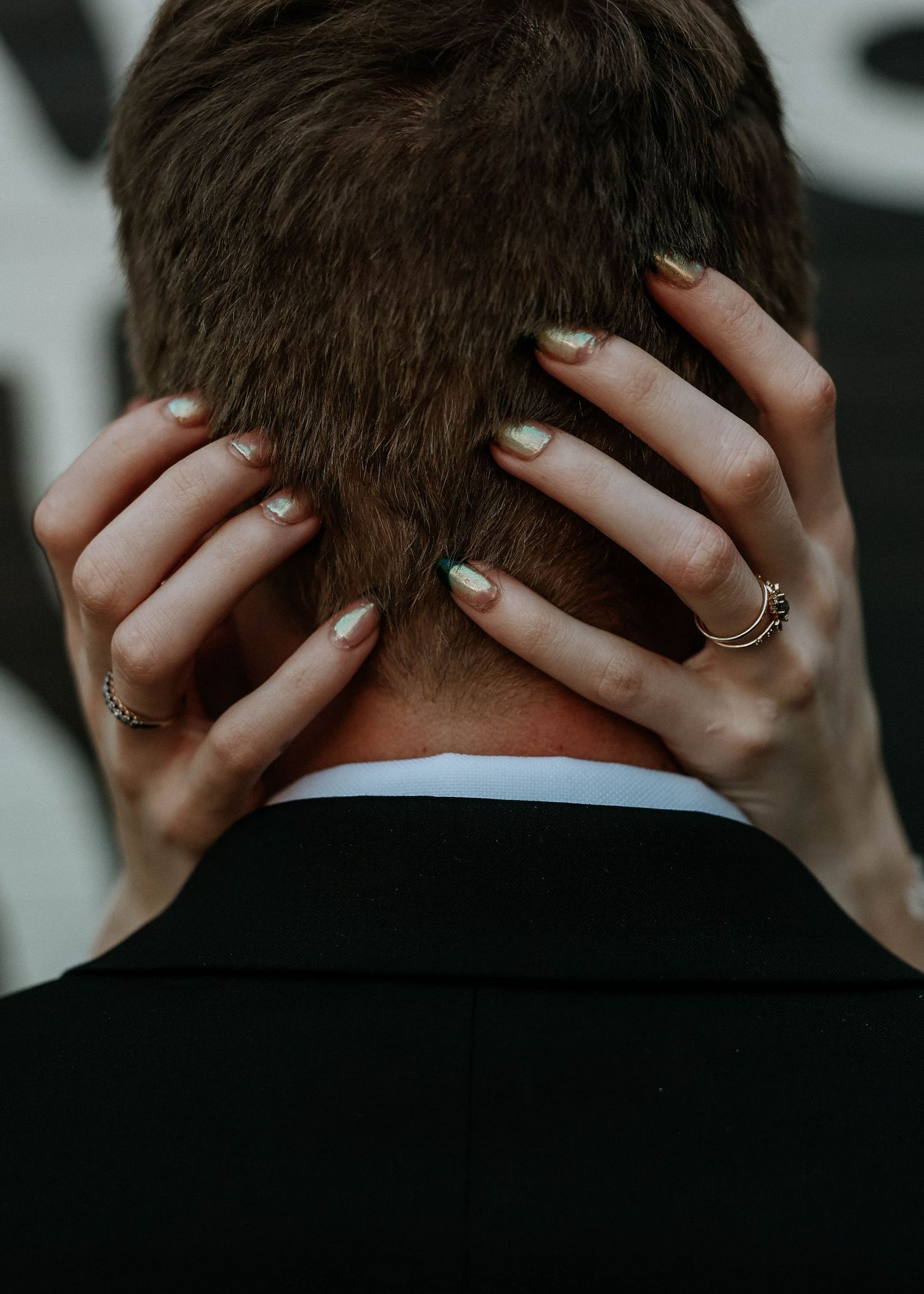 Woman's hands with gold rings and sage nail polish cradling the back of a man's head in a black suit jacket.