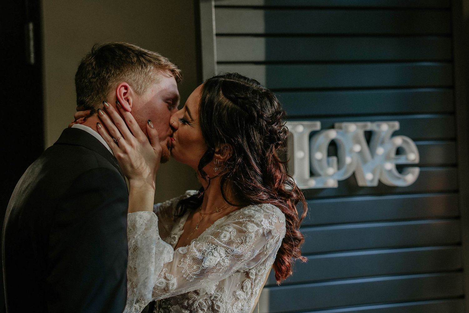 A couple shares a romantic kiss in front of an illuminated Love sign at their wedding reception.