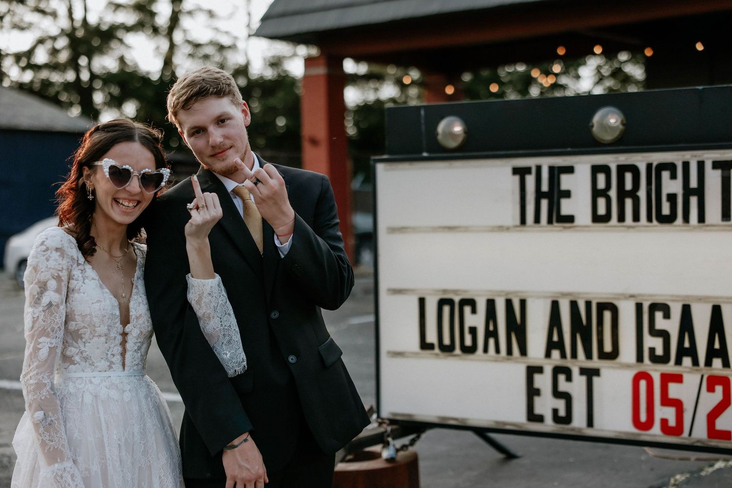 Newlyweds Logan and Isaac pose playfully beside a marquee sign celebrating their wedding date EST 05/2.
