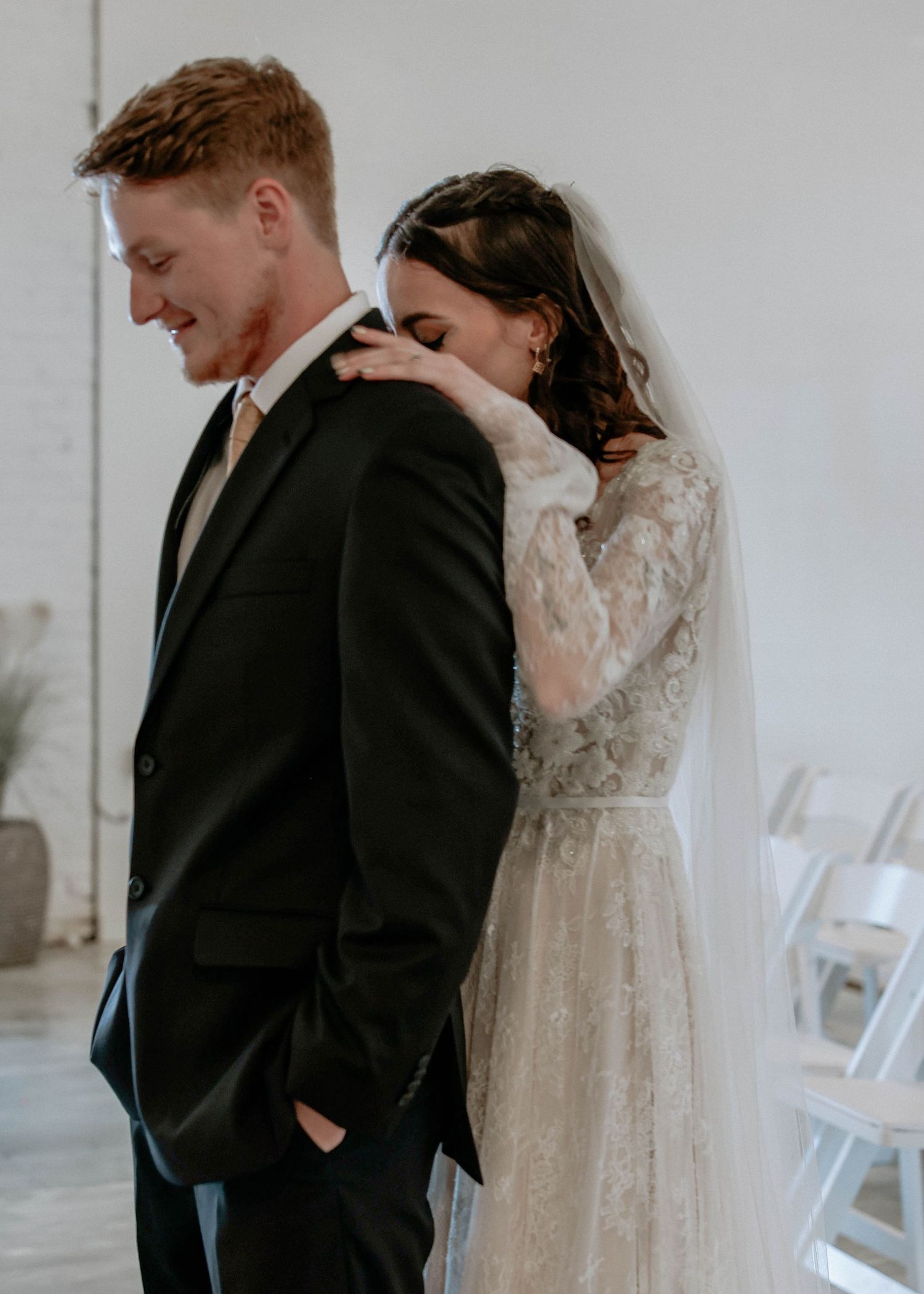 Bride in lace gown rests head on groom's shoulder during emotional first look at wedding ceremony.