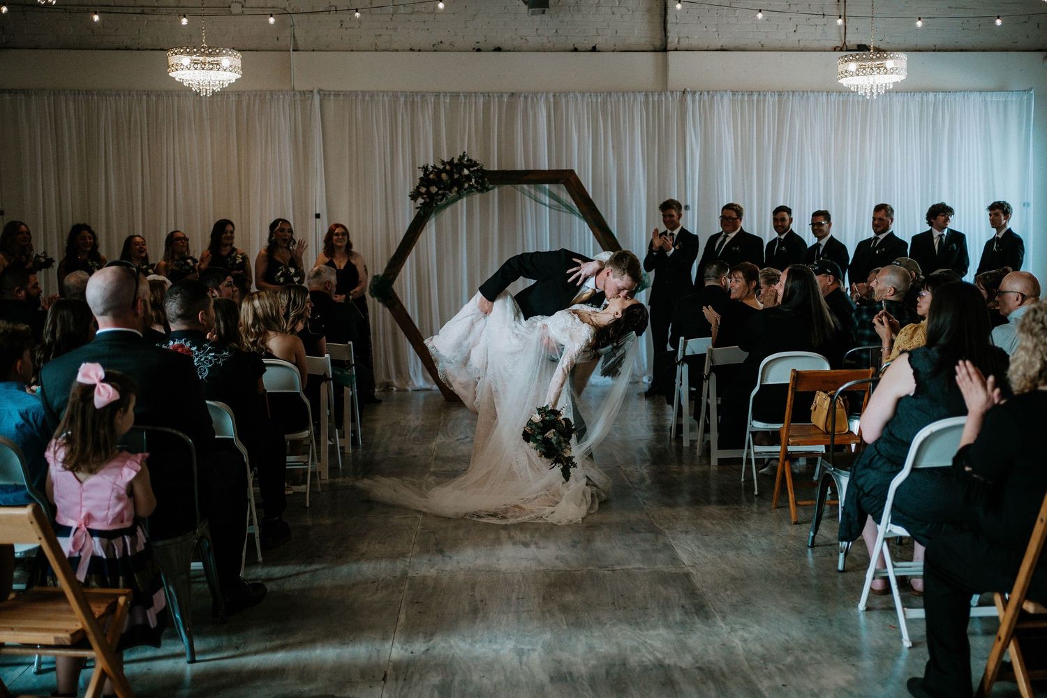 Groom dips bride for a kiss at indoor wedding ceremony with hexagonal arch and seated guests.