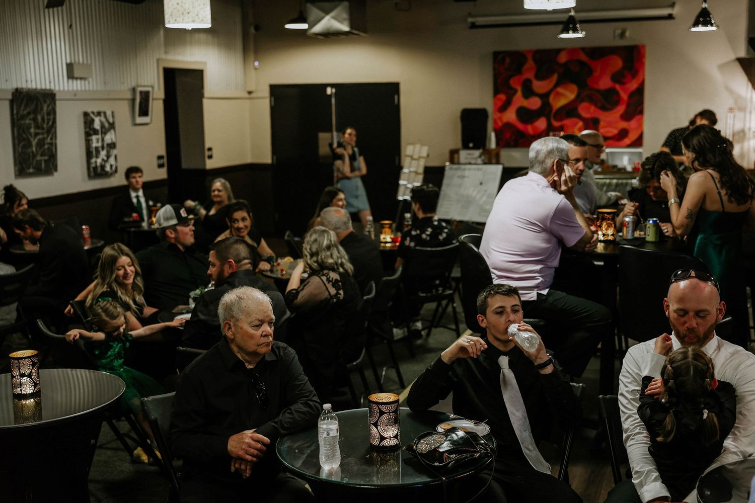 Guests socializing at a dimly lit event venue with black tables, drinks, and artwork on the walls.