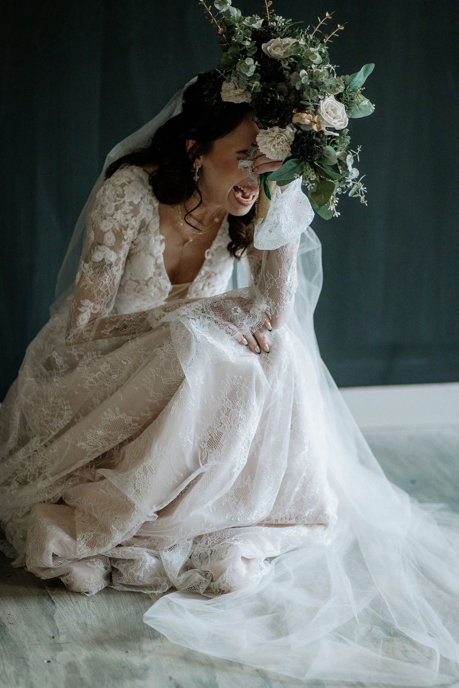 Bride in lace wedding gown sitting on floor laughing while holding a lush green and ivory floral bouquet.