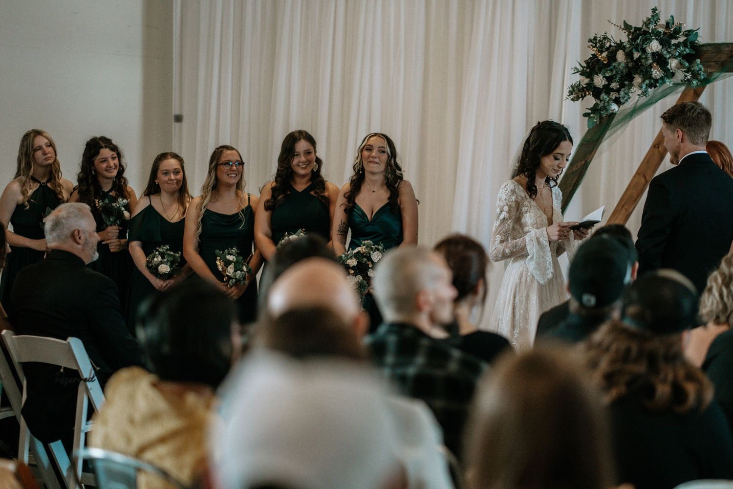 Bridesmaids in emerald green dresses stand at a wedding ceremony altar with floral arch and bride in lace gown.