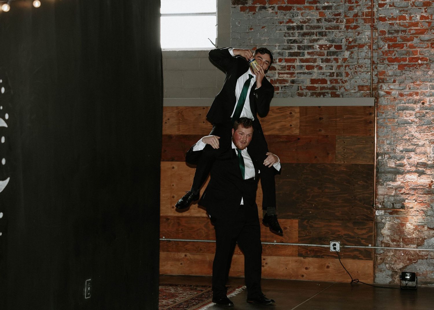Two groomsmen in black tuxedos playfully pose at a wedding reception in a rustic brick venue.