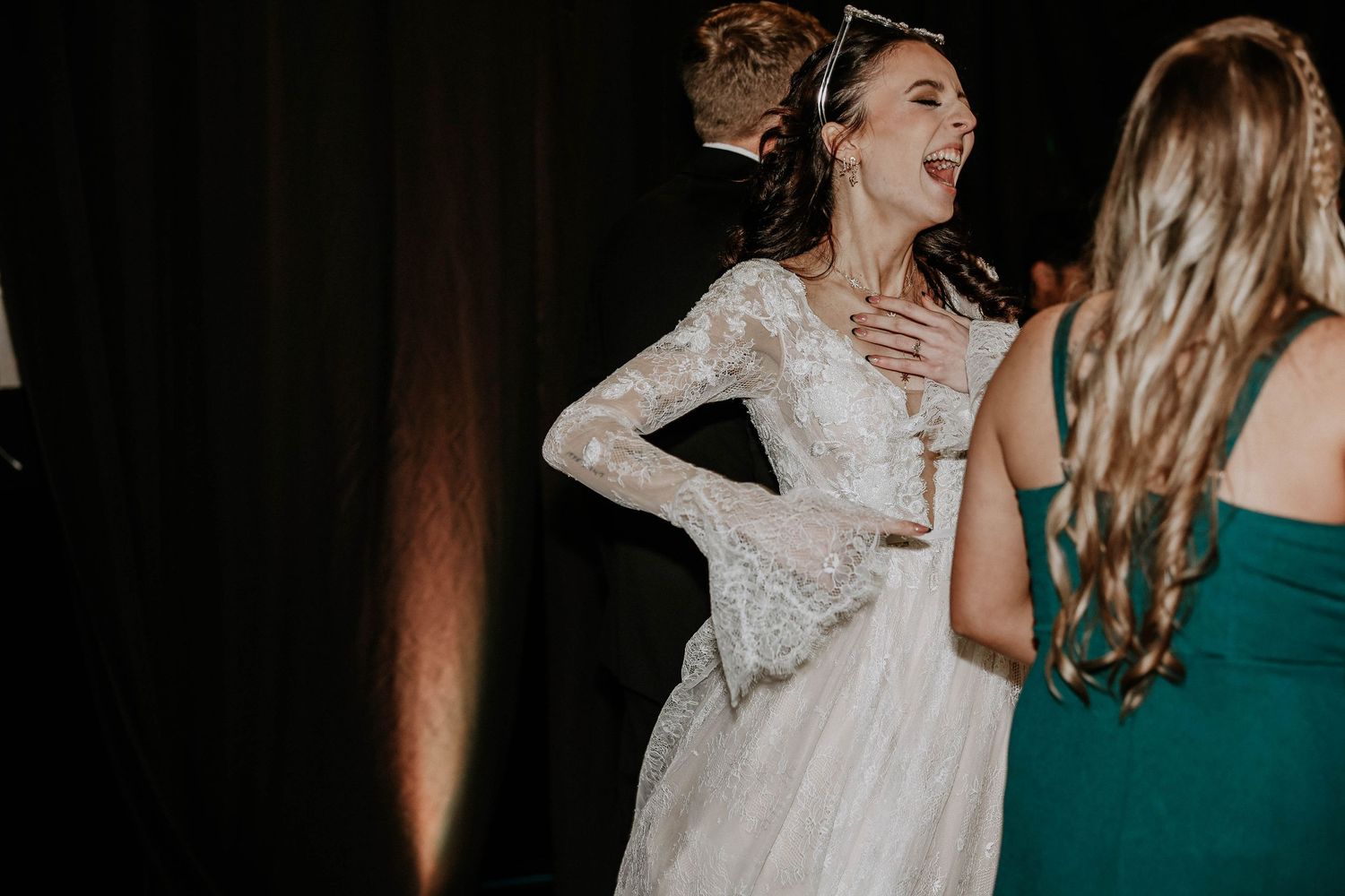 Bride in lace long-sleeve wedding gown laughing joyfully with guests at elegant wedding reception.