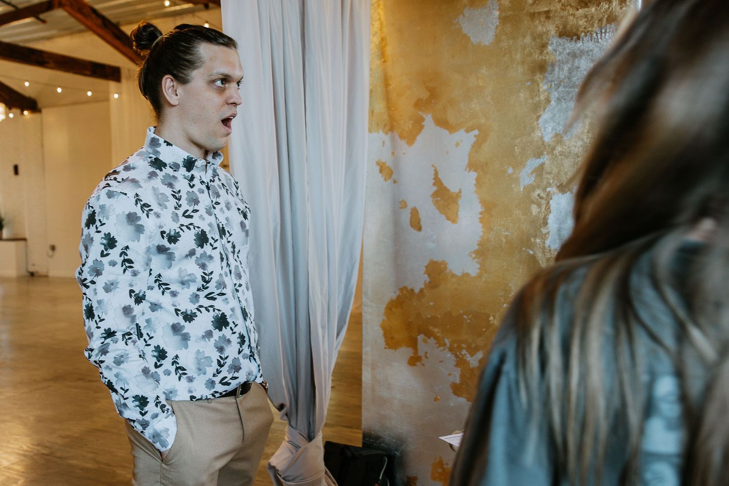 Man in floral shirt standing near sheer curtains in a rustic venue.