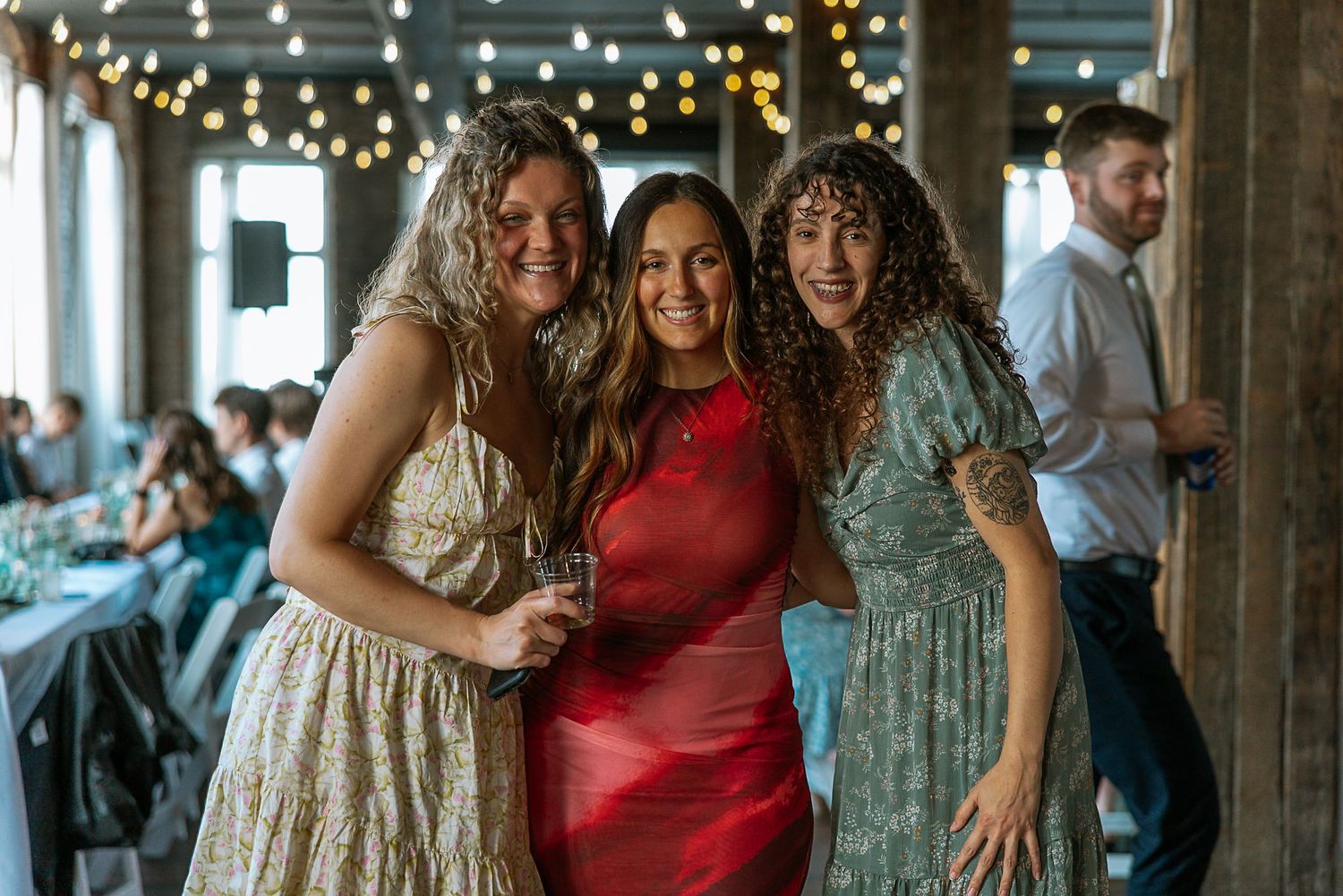 Three smiling women posing together at a wedding reception with string lights.