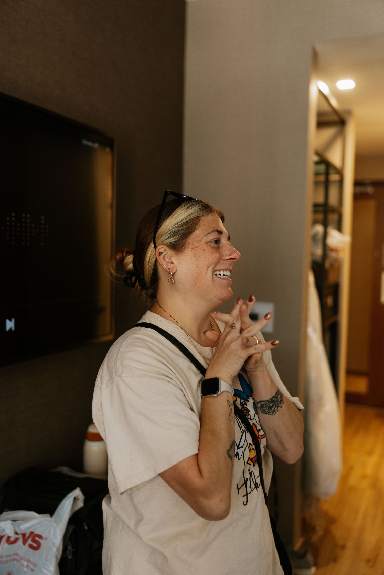Blonde woman laughing joyfully in a hotel room wearing a white t-shirt and smartwatch.