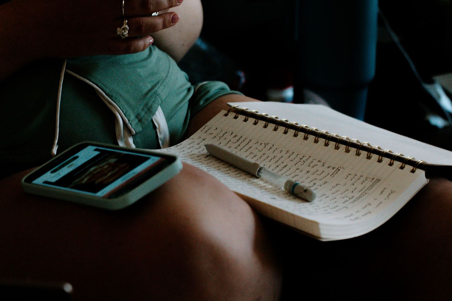 Bride in green pajamas reviews handwritten wedding vows in a spiral notebook beside her smartphone.