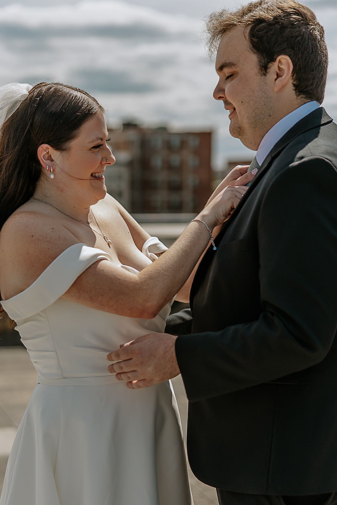 Bride in off-shoulder gown smiles at groom while adjusting his tie on a rooftop wedding day.