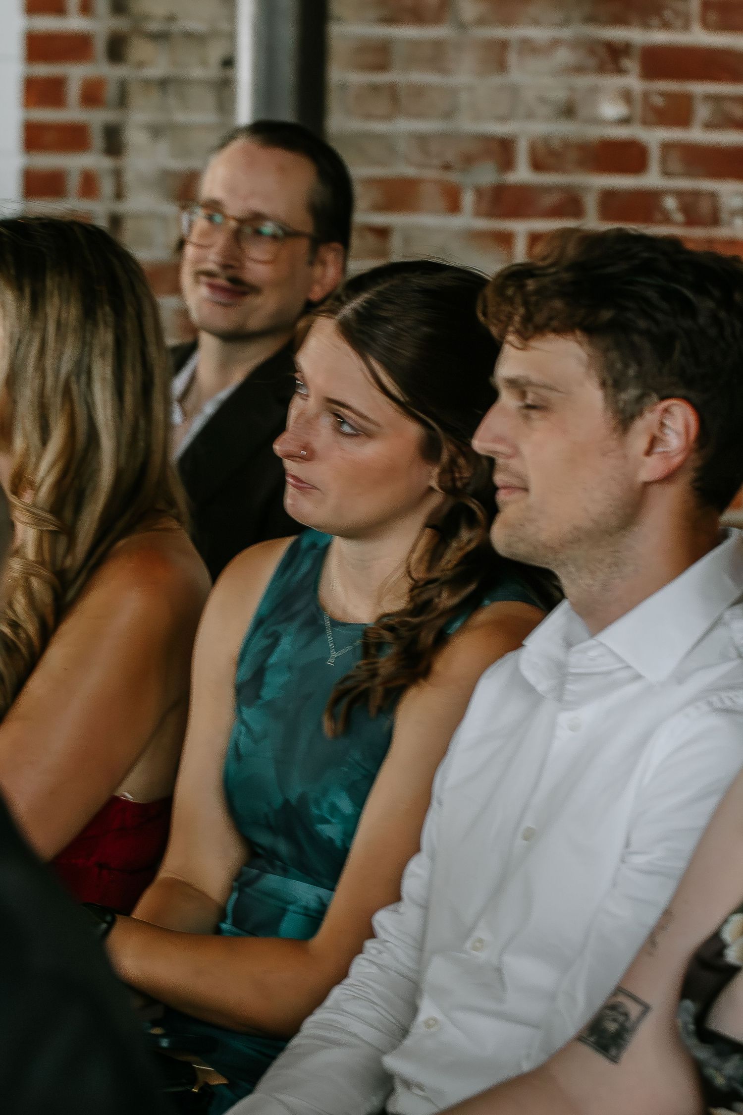 Emotional wedding guests watching ceremony in rustic brick venue with string lights.