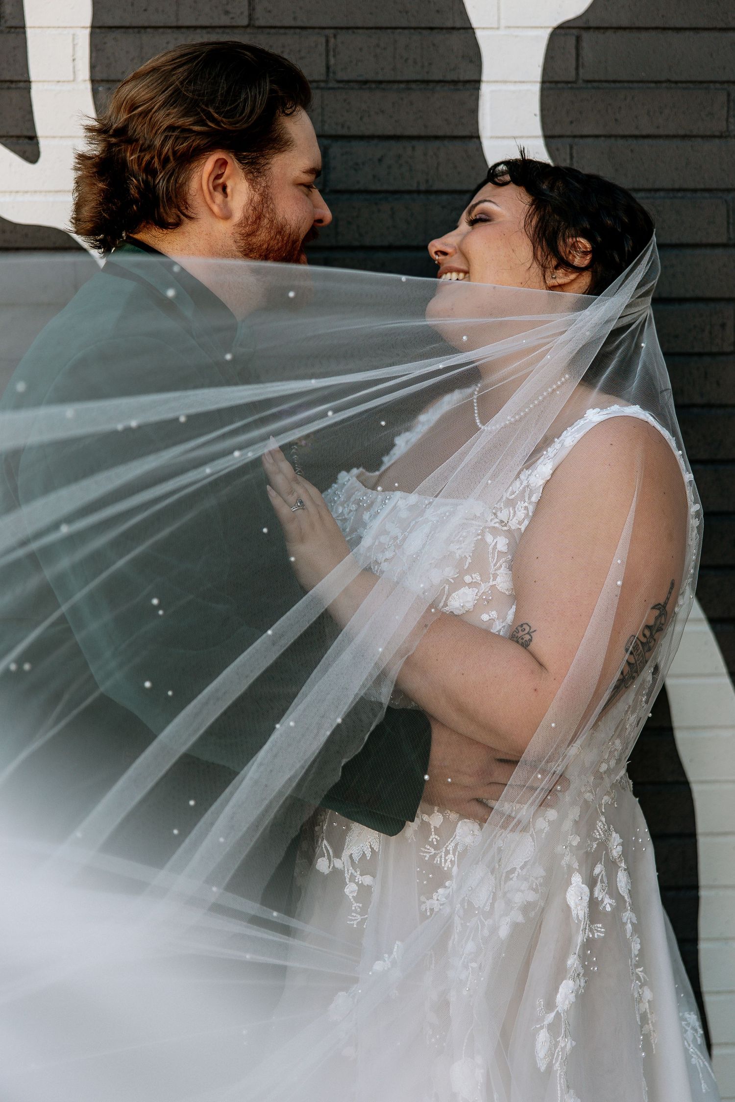 Bride and groom sharing a joyful moment together, wrapped in a flowing pearl-dotted wedding veil outdoors.