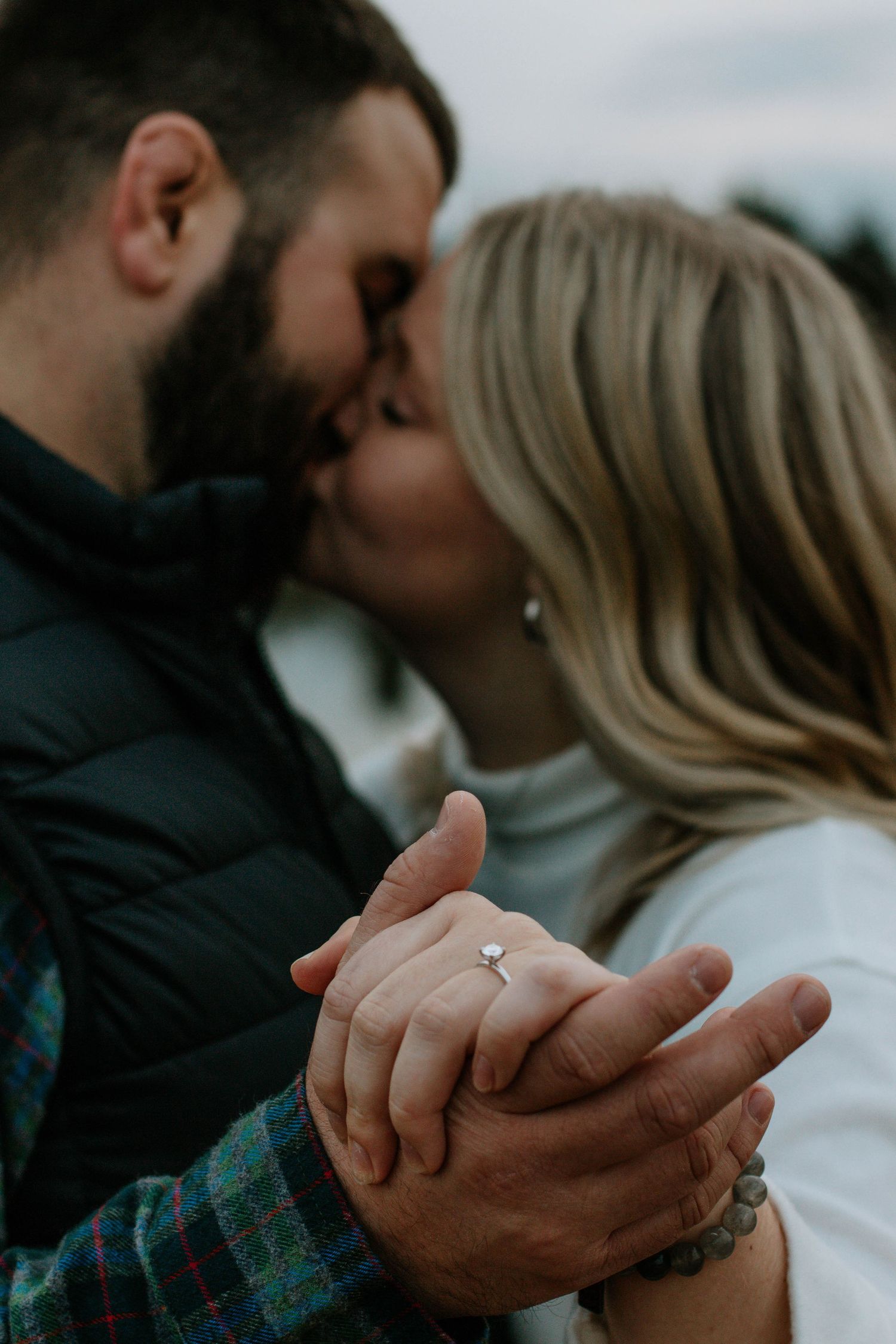 Couple sharing a kiss while holding hands, showcasing an engagement ring with bokeh background in Dayton, Ohio
