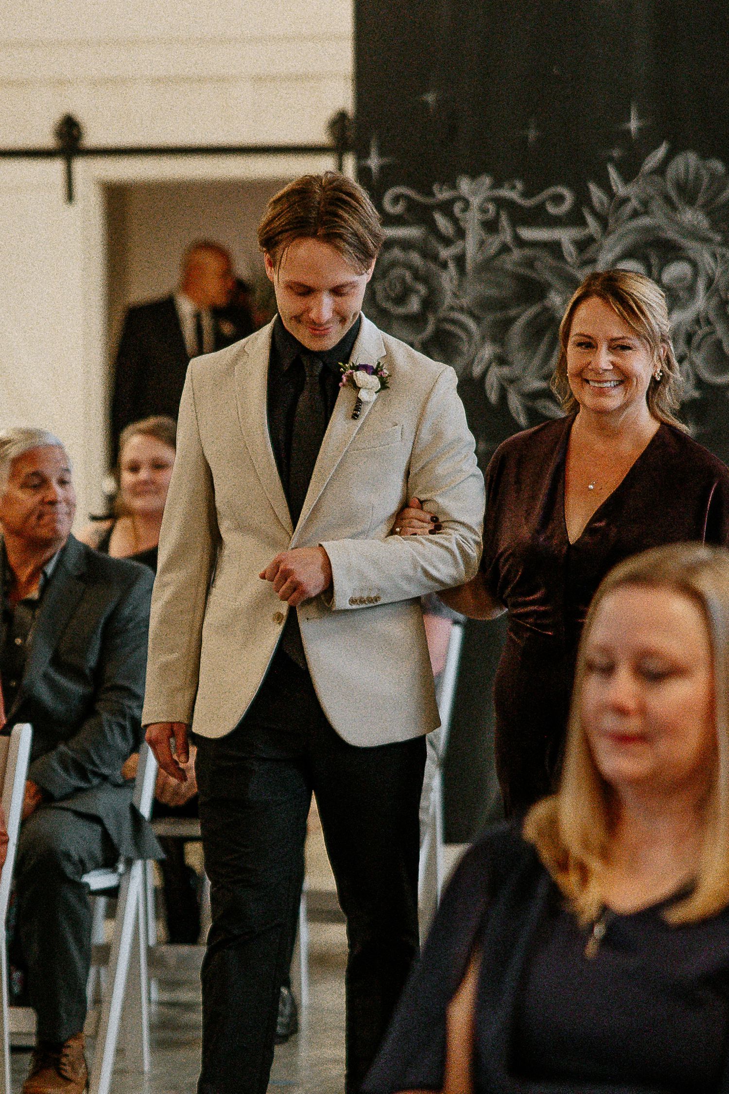 Groom in ivory blazer escorts smiling woman down wedding ceremony aisle.