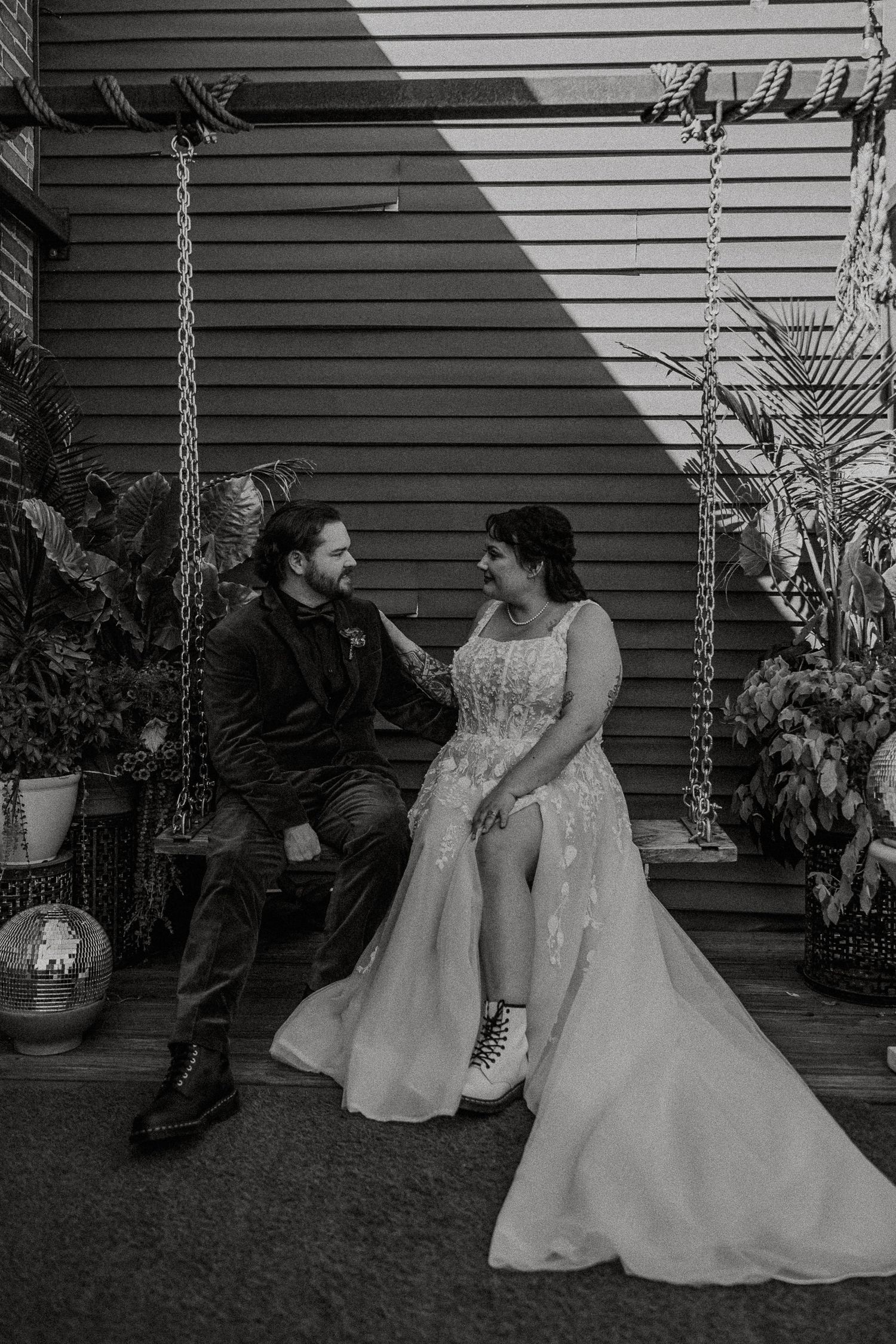 Black and white photo of bride and groom sitting on outdoor swing, smiling at each other.