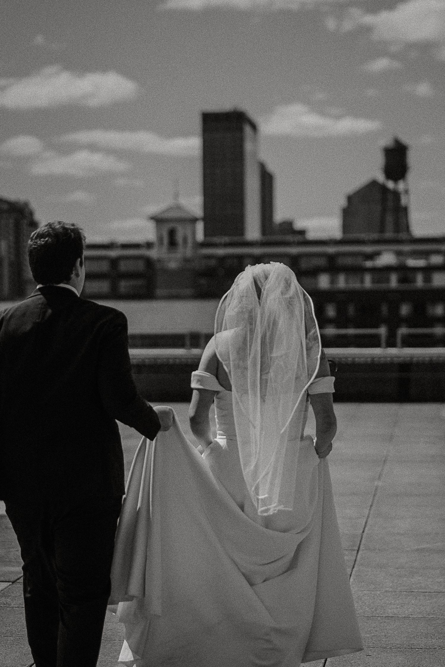 Black and white photo of newlyweds walking hand in hand on rooftop with city skyline behind them.