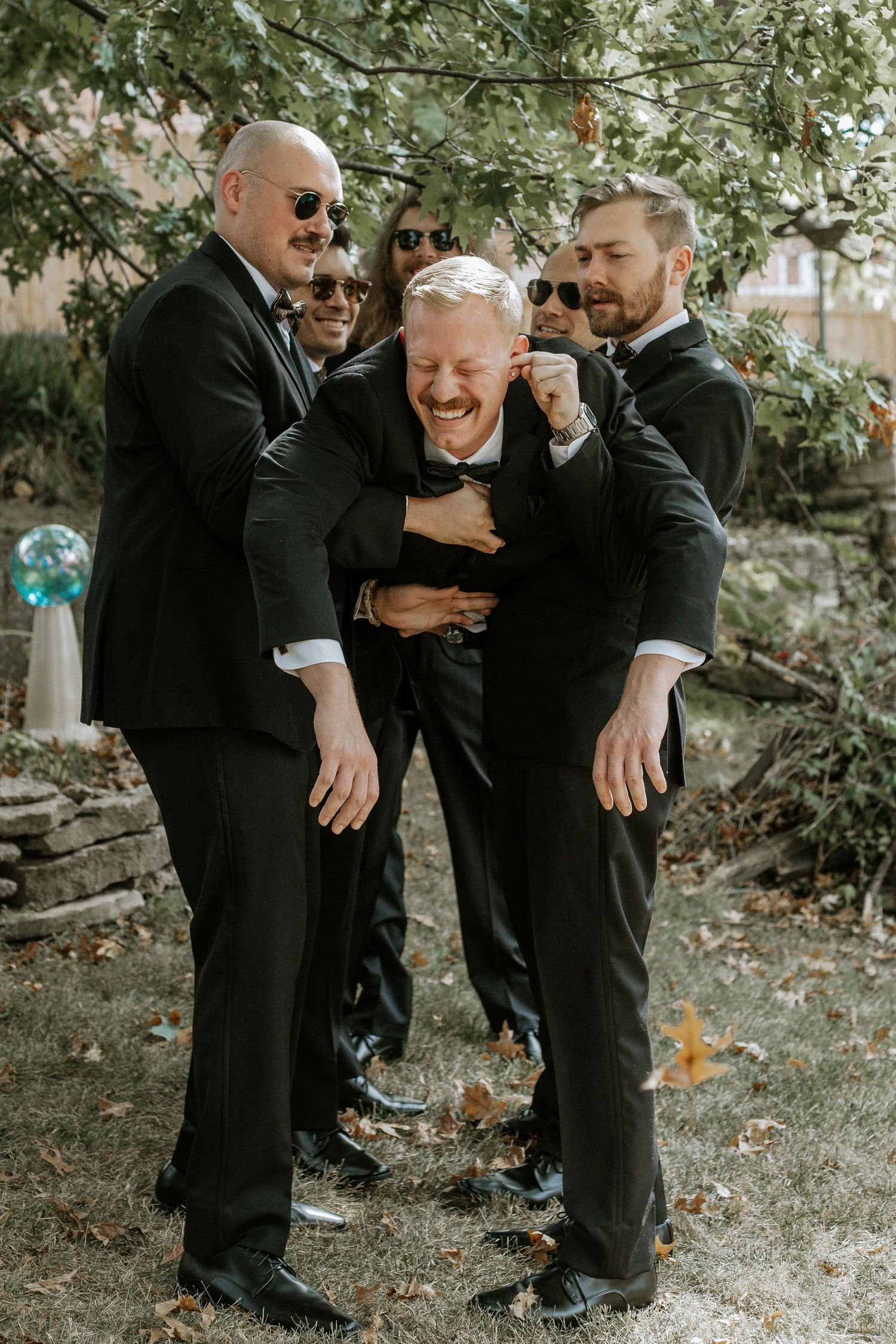Groomsmen in black tuxedos laugh and playfully embrace the groom outdoors under a tree in Cincinnati, Ohio