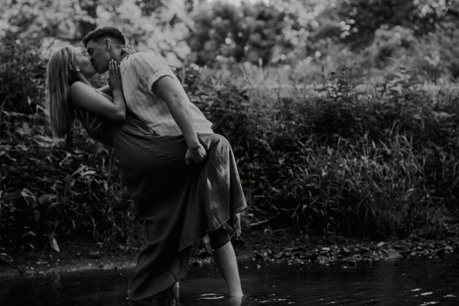 Black and white photo of a couple sharing a romantic kiss near a serene creek surrounded by lush greenery at Eden Park in Cincinnati, Ohio
