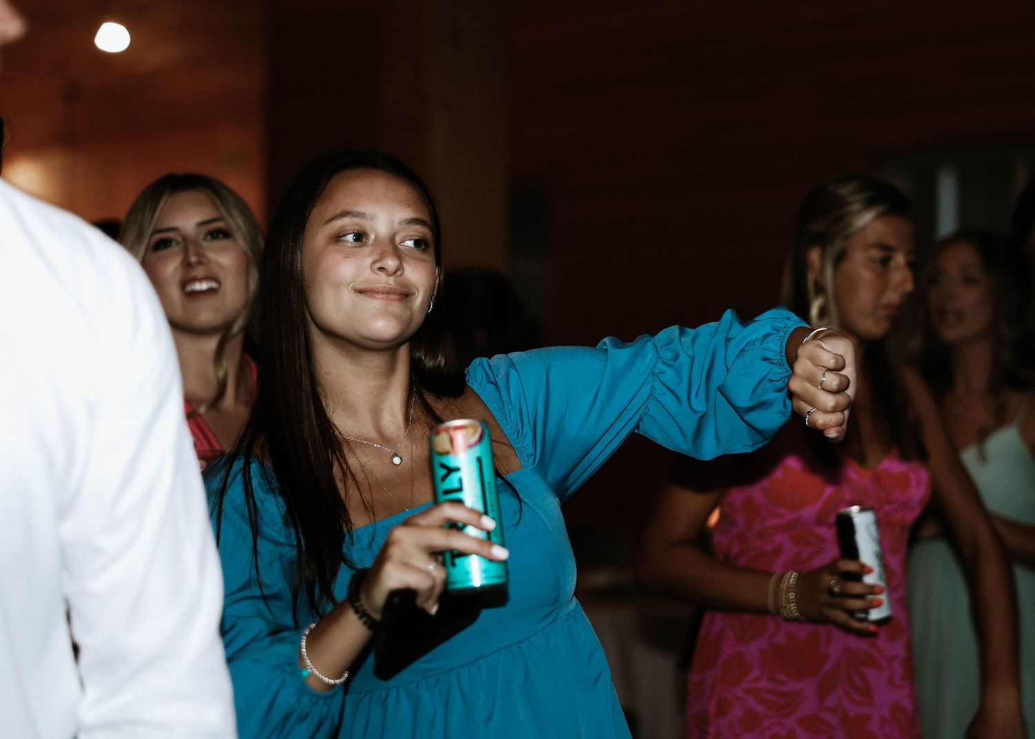Woman in teal dress dancing at party, holding drink cans, with guests in background.